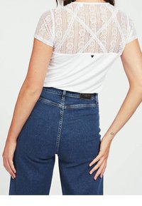 Hvid t-shirt med blondedetalje bagpå, korte ærmer, kombineret med højtaljede blå jeans. Jeansene har standard baglomme og mærkeetiket.