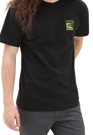 Mand iført en sort Vans t-shirt med et lille grønt geometrisk logo på venstre bryst, parret med mørkegrå bukser.