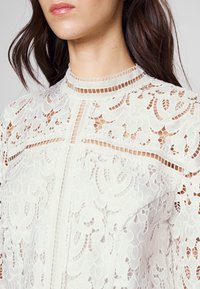 Krajková blůza v krémové barvě s propracovanými květinovými vzory, vysokým výstřihem a průhlednými detaily, s přiléhavým střihem.