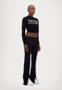Suéter cropped preto com o logótipo 'Versace Jeans Couture', combinado com calças pretas ajustadas com aberturas laterais e bainhas rendadas. Ténis brancos.