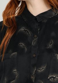 Svart skjorta med texturerad tyg, prydd med guldfladdermusmönster, en spetskrage och bruna knappar framtill. Två bröstfickor synliga.