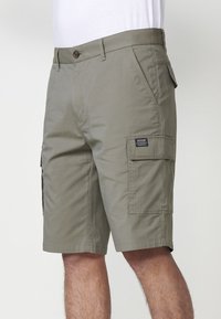 Uomo che indossa pantaloni cargo beige con tasche a bottone e a patta, in piedi contro uno sfondo chiaro e semplice.