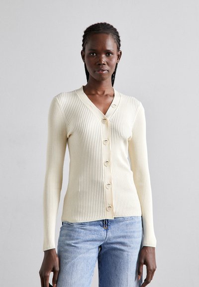 Cream ribbed cardigan with a V-neck, featuring six matching buttons down the front and long sleeves. Worn over blue jeans.