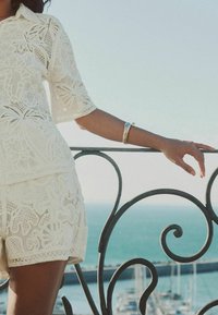 Femme portant une tenue en dentelle blanche avec un bracelet repose son bras sur une rambarde en métal noir courbée surplombant la marina et l'océan.