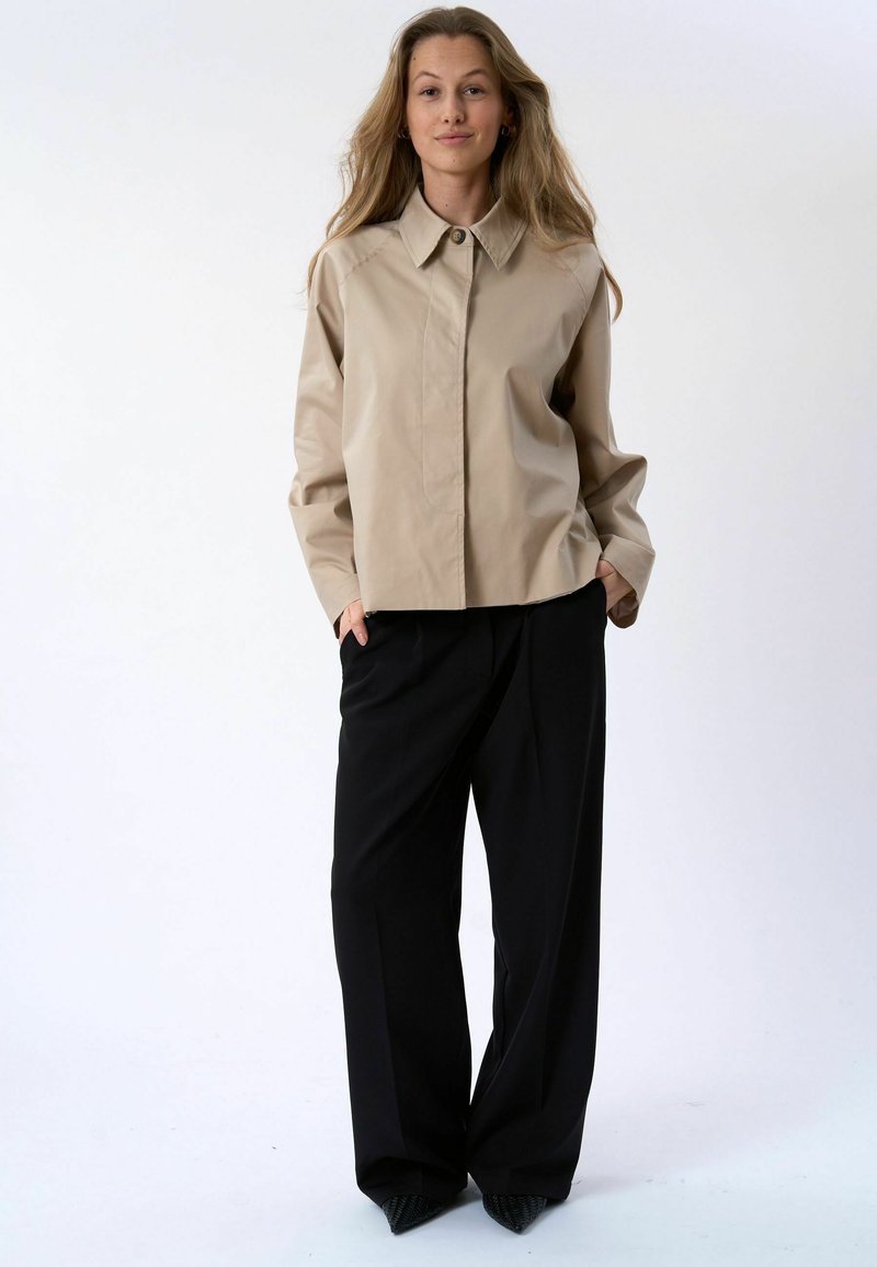 Femme debout, les mains dans les poches, portant une chemise beige boutonnée et un pantalon noir à jambes larges sur un fond blanc uni.