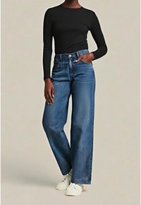 Personne portant un t-shirt noir à manches longues, un jean bleu taille haute à jambes larges, des chaussures blanches, debout avec une main dans la poche.