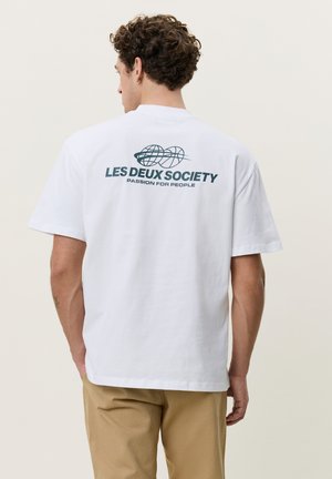 Mand med krøllet hår iført hvid T-shirt med globuslogo og teksten "LES DEUX SOCIETY Passion for People," iført beige bukser, stående med ryggen til.