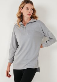 Svetlo siva zip-up pulover s polovičnim ovratnikom, dolgimi rokavi in stranskimi razpoki. Mehka, gladka tkanina z sproščenim krojem.