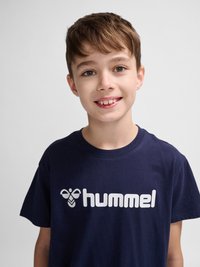 Uśmiechnięty chłopiec z krótkimi brązowymi włosami, ubrany w granatowy t-shirt marki Hummel na jednolitym jasnoszarym tle.