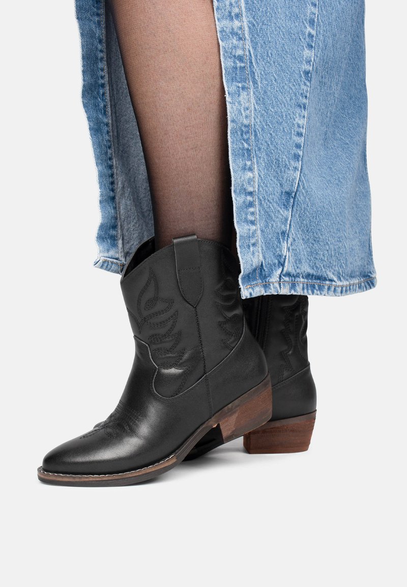 Botas de tobillo de cuero negro con punta afilada, costuras decorativas y tacón cuadrado marrón, combinadas con una falda de mezclilla.