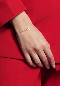 Złota bransoletka łańcuszkowa z zawieszką w kształcie serca, noszona na dłoni na tle czerwonej tkaniny. Subtelny blask, delikatny design.