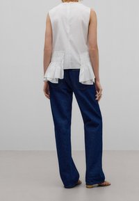 Haut sans manches blanc avec des accents à volants sur les côtés, associé à un jean à jambes larges bleu foncé. Tissu lisse, design minimaliste et fermeture éclair au dos.