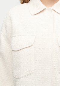 Kremowa kurtka o aksamitnej fakturze z ostro zakończonym kołnierzem i dwoma dużymi kieszeniami na piersi z klapami i lamówkami.