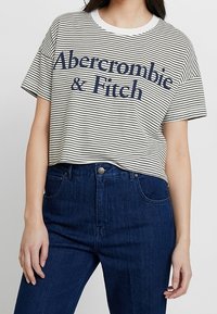 Pruhované tričko s krátkým rukávem v krémové a námořnické barvě s nápisem "Abercrombie & Fitch". Materiál z bavlny, kulatý výstřih, volný střih. V kombinaci s tmavě modrými džíny.