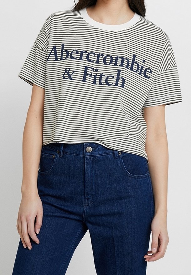 Pruhované tričko s krátkým rukávem v krémové a námořnické barvě s nápisem "Abercrombie & Fitch". Materiál z bavlny, kulatý výstřih, volný střih. V kombinaci s tmavě modrými džíny.