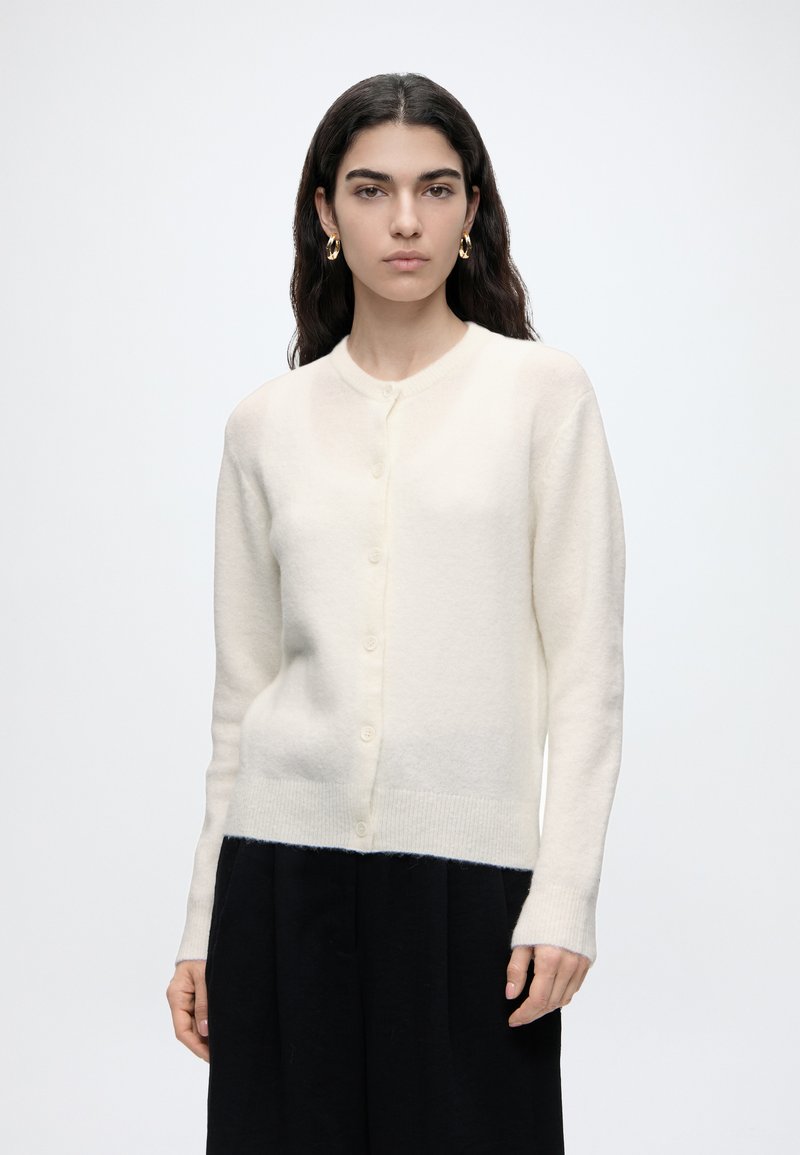 Jeune femme aux longs cheveux foncés portant un cardigan blanc boutonné, un pantalon noir et des boucles d’oreilles créoles dorées sur un fond clair uni.