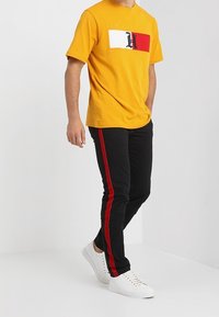 Gul t-shirt med kort ärm och ett horisontellt mönster av vit och röd randig logotyp, tillsammans med svarta byxor med röda sidostripes och vita sneakers.