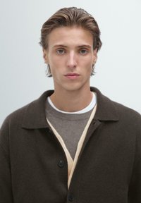 Mladý muž se světle hnědými vlasy, oblečený v vrstevnatých hnědých a béžových svetrech a tmavě hnědém zapínacím kabátu, dívající se vpřed proti jednobarevnému pozadí.