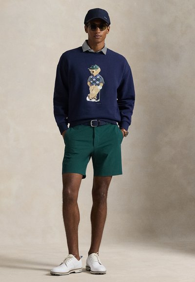 Μπλε σκούρο φούτερ με γραφικό αρκούδας, πράσινα σορτς, λευκά παπούτσια και μπλε καπέλο. Υφή υφάσματος με ρίγες στο γιακά. Μινιμαλιστικός σχεδιασμός.