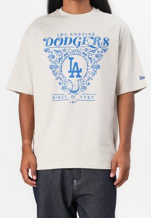 Personne portant un T-shirt beige clair avec le logo bleu des Los Angeles Dodgers, un design floral, un graphisme de baseball et le texte depuis 1958.