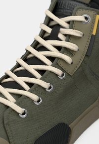 Πράσινο υφασμάτινο παπούτσι τύπου high-top με μπεζ κορδόνια, μαύρες λεπτομέρειες, υφή επιφάνειας και ασημί οπές για τα κορδόνια.