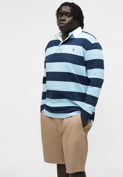 Άνδρας με σκουρόχρωμο δέρμα και dreadlocks φορά μπλε μπλουζάκι πόλο με ριγέ μακριά μανίκια και μπεζ σορτς, στέκεται μπροστά σε απλό ανοιχτό γκρι φόντο.