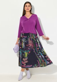 Lila, langärmliges Oberteil mit V-Ausschnitt, kombiniert mit einem plissierten, mehrfarbigen Rock mit Blumenmuster und einer helllila Clutch.