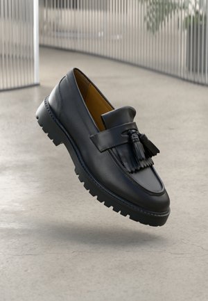 Mocassin à pampilles en cuir noir avec semelle crantée épaisse flottant au-dessus d'un sol gris clair dans un décor intérieur moderne.