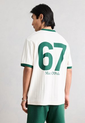 Άνδρας με λευκό κοντομάνικο πουκάμισο με πράσινες λεπτομέρειες και μεγάλο πράσινο αριθμό 67 με το κείμενο «Marc O’Polo» στο πίσω μέρος, συνδυασμένο με πράσινα σορτς.