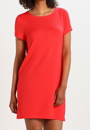 Frau trägt ein rotes, knielanges Kleid mit kurzen Ärmeln und rundem Ausschnitt, steht vor einem einfarbigen hellen Hintergrund.