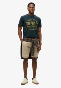Mænds mørkeblå t-shirt med gul tekst, beige shorts og beige sneakers. En grå sweatshirt bundet om livet giver kontrast.
