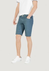 Bermudas azules confeccionadas en tela ligera, con cierre frontal de botón, bolsillos laterales y un corte entallado. Combinadas con zapatillas blancas.
