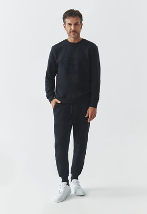 Sudadera azul marino con un gráfico de oso, combinada con pantalones jogger a juego. Presenta puños y cintura acanalados, llevada con zapatillas blancas.
