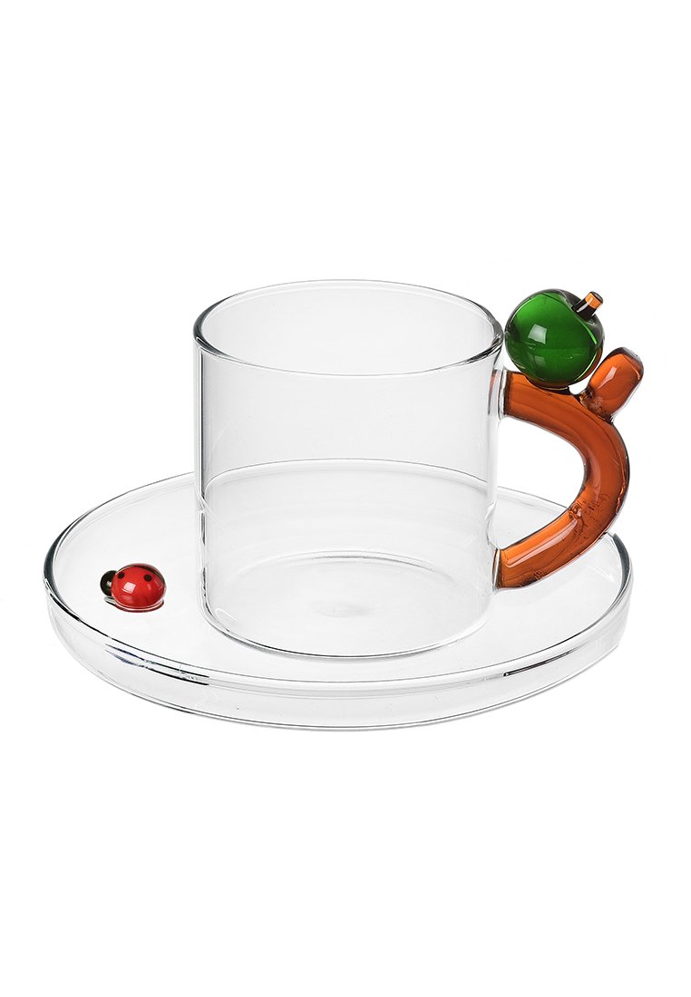 Szklanka z pomarańczowym uchwytem ozdobionym zielonym okrągłym akcentem, podana na przejrzystym spodku z czerwonym szczegółem w kształcie biedronki.