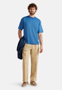 T-shirt di cotone blu con maniche corte, pantaloni di lino beige e scarpe marroni. Una giacca scura è appoggiata sul braccio sinistro.