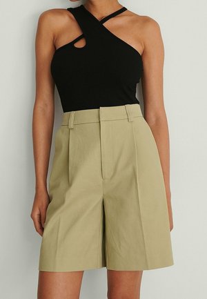 Frau trägt beige, hoch taillierte, maßgeschneiderte Shorts und ein schwarzes asymmetrisches Neckholder-Top mit Ausschnittdetail an der Schulter.