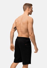 Shorts atletici neri con vita elastica, dotati di una tasca sul retro e texture liscia. Indossati da un modello maschile visto da dietro.