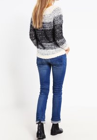 Strickpullover mit einem dicken, strukturierten Muster in schwarz-weißen Streifen, kombiniert mit schmalen blauen Jeans und schwarzen Stiefeletten.