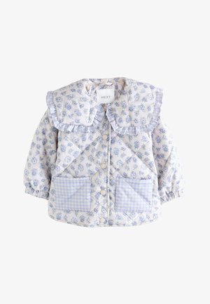 Lachende peuter met krullend haar, een blauwe bloemige jas met grote kraag, blauwe spijkerbroek en roze sneakers tegen een beige achtergrond.