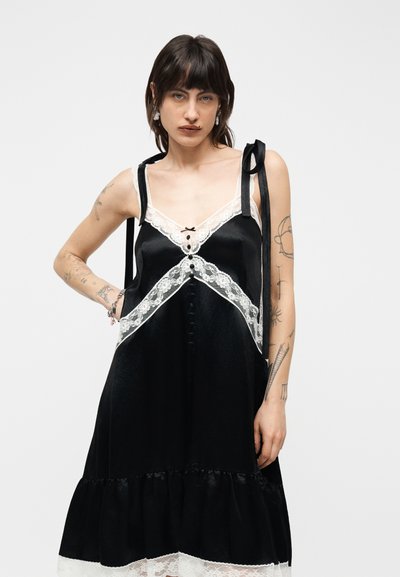 Νεαρή γυναίκα με τατουάζ φοράει μαύρο σατέν φόρεμα με λευκή δαντέλα στο τελείωμα και κορδέλες στους ώμους, στέκεται μπροστά σε απλό φόντο.