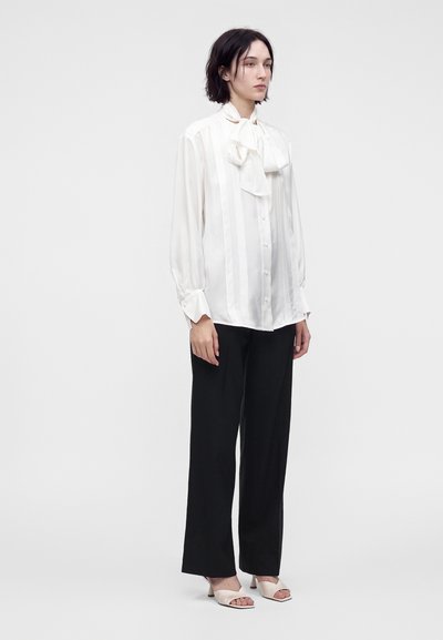 Femme debout portant un chemisier blanc à manches longues avec un col à nœud papillon, un pantalon noir à jambes larges et des sandales à talons ouvertes beiges.