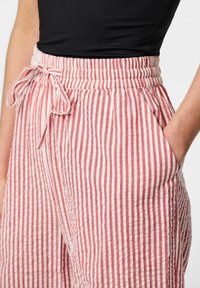 Gestreepte, lichte broek in rood en crème met een elastische tailleband en koordje. Beschikt over zijzakken en een gestructureerde stof.