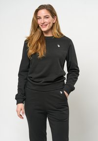 Svart sweatshirt- och byxset i bomull. Lång ärm, ribbade mudd, rund halsringning, med en liten vit logotyp. Slät yta.
