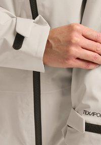 Veste imperméable gris clair avec fermeture éclair et accents noirs. Dispose de poignets ajustables avec Velcro et de la marque "TEXPOR" sur la manche.