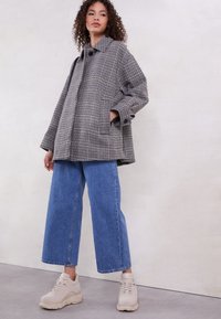 Oversized grijze coat met een haakjesmotief en een kraaghals, gecombineerd met een wijde spijkerbroek in blauw en lichtbeige sneakers.
