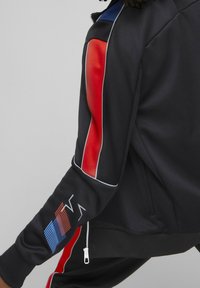 Černá sportovní bunda s červenými a modrými akcenty, obsahující kapsu na zip, kontrastní prošívání a texturovanou část na rukávu.