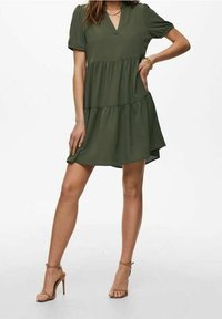 Robe olive verte à volants avec manches courtes bouffantes, encolure en V et ourlet fluide. Associée à des talons à brides marron clair et un bracelet. Texture de tissu lisse.