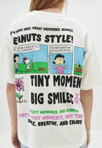 Hvid bomulds T-shirt med farverige grafikker i tegneseriestil med Snoopy og Lucy, store tekstsætninger og en blomsterillustration.