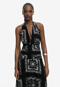 Schwarzes Halsbandkleid mit weißem Bandana-Muster, tiefem V-Ausschnitt, gerafft in der Taille und fließendem Rock. Silberner Armschmuck als Akzent.