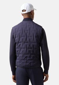 Navy quilted vest with a textured finish, side mesh panel, and ribbed collar. Lightweight and fitted design, paired with a long-sleeve top.